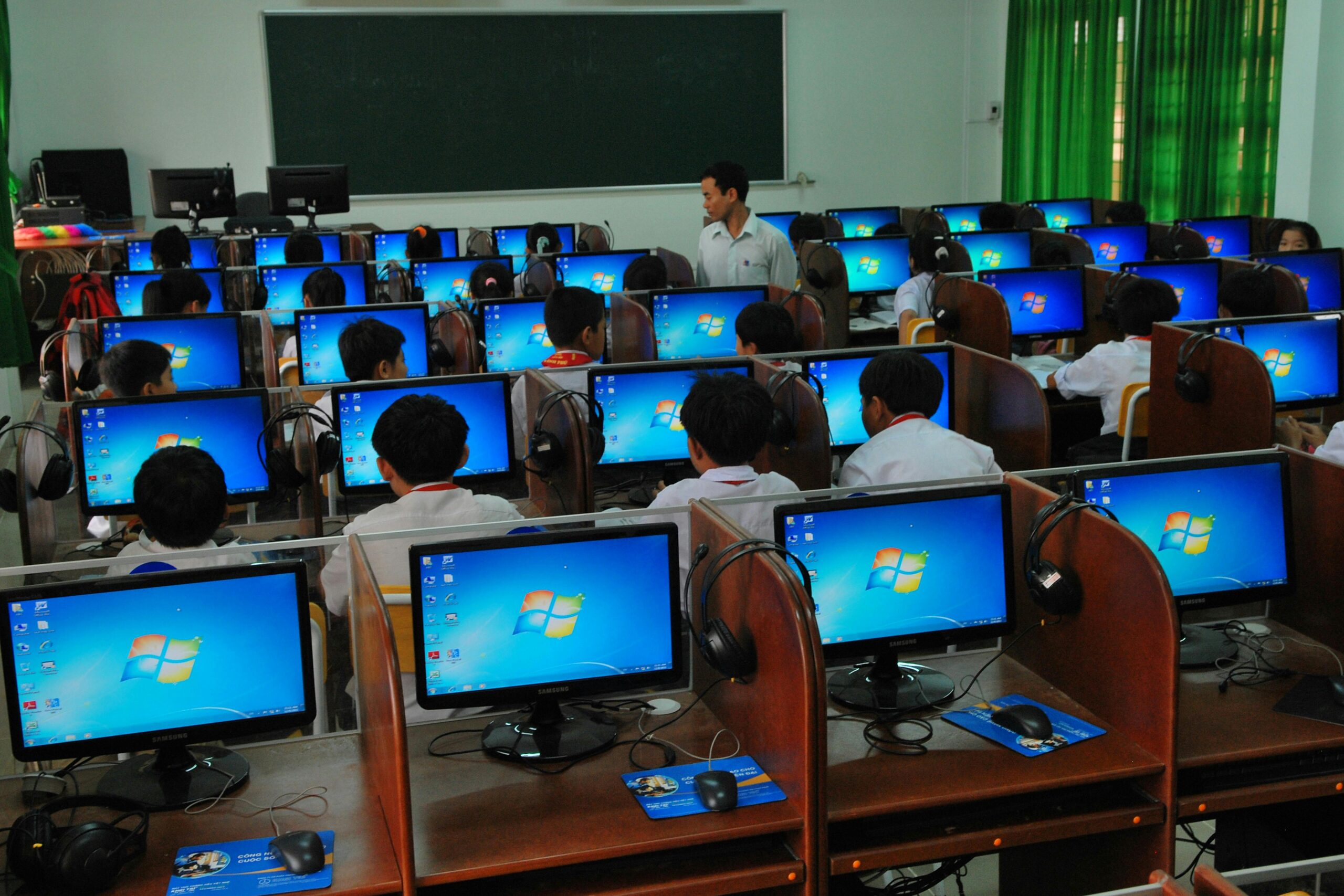 Students learning in a computer lab with multiple monitors and a teacher instructing.